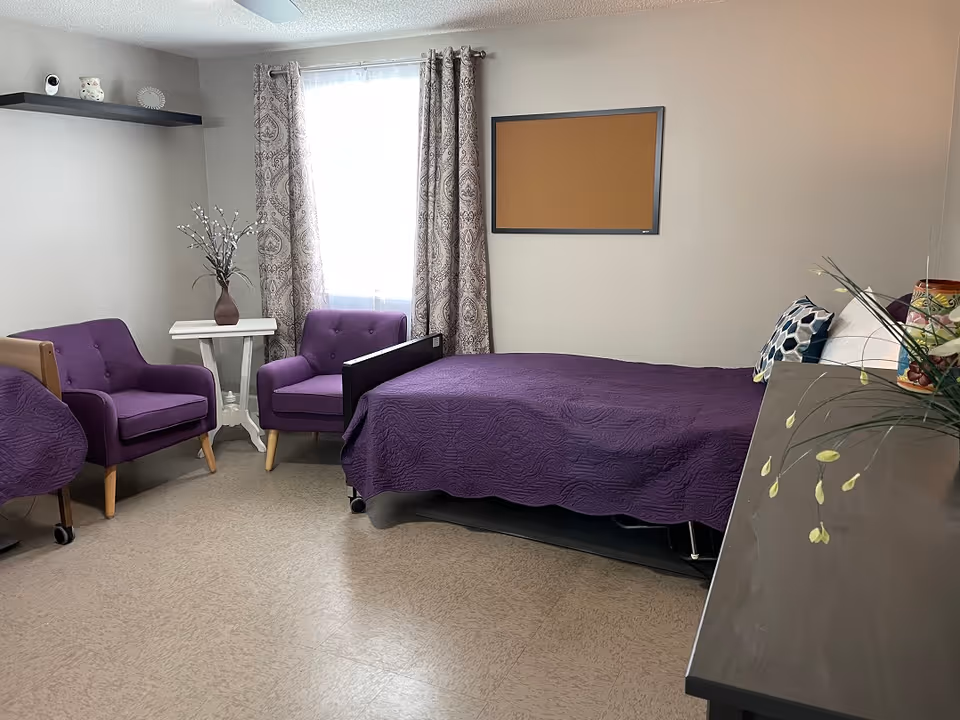 A cozy bedroom in a senior living facility with a single bed covered in a purple quilt, two purple armchairs, a small white side table with a vase of flowers, patterned curtains on the window, a corkboard on the wall, and a dresser with decorative items and plants.
