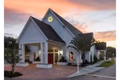 Front entrance of a white, chapel-like senior living building with a peaked roof and covered porte-cochère at sunset.