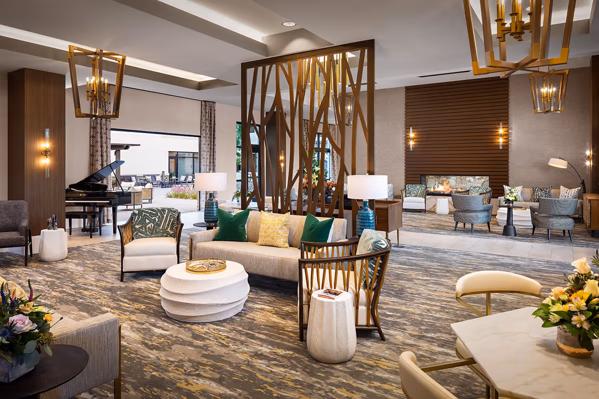 Spacious, well-lit senior living lounge with modern seating, a fireplace, piano, decorative room divider, and tables.