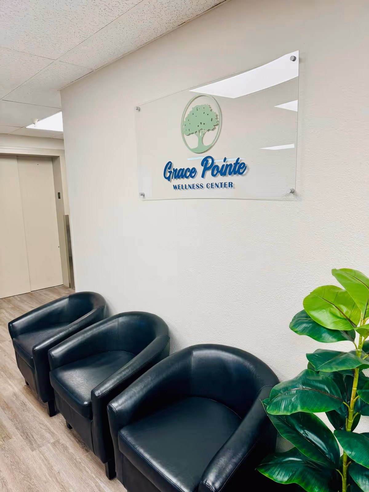 Waiting room with three black chairs, a potted plant, and a wall sign that reads "Grace Pointe Wellness Center".