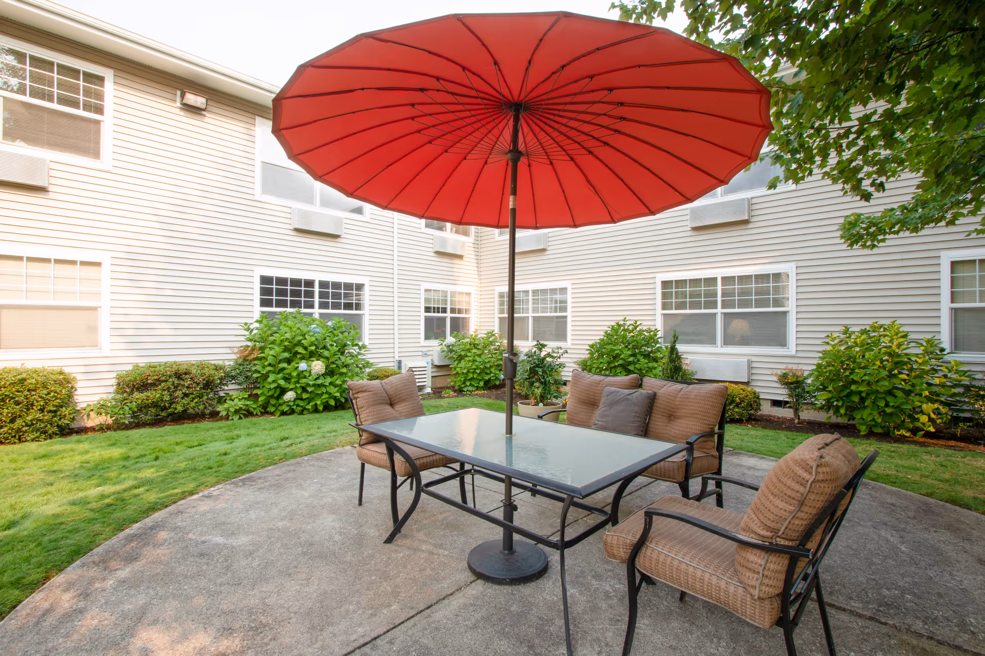 Outdoor patio area with a glass-top table, four cushioned chairs, and a large red umbrella providing shade. The patio is surrounded by green grass, bushes, and the exterior walls of a building with multiple windows.