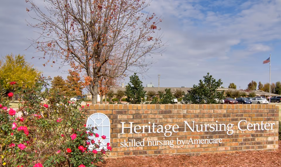 Brick sign for Heritage Nursing Center with the text 'skilled nursing by Americare' surrounded by bushes with pink flowers and trees, with a parking lot and American flag in the background under a partly cloudy sky.