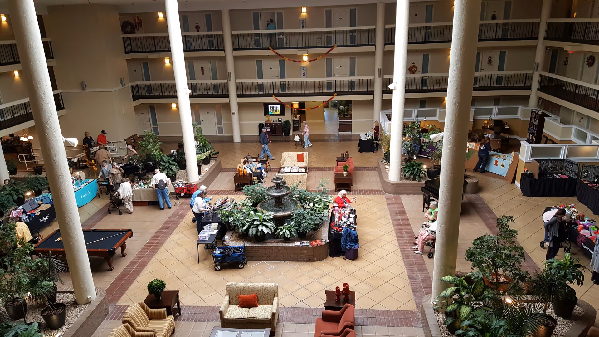 Multi-story atrium in a senior living facility with a central fountain, seating areas, plants, and residents gathered around tables and booths.