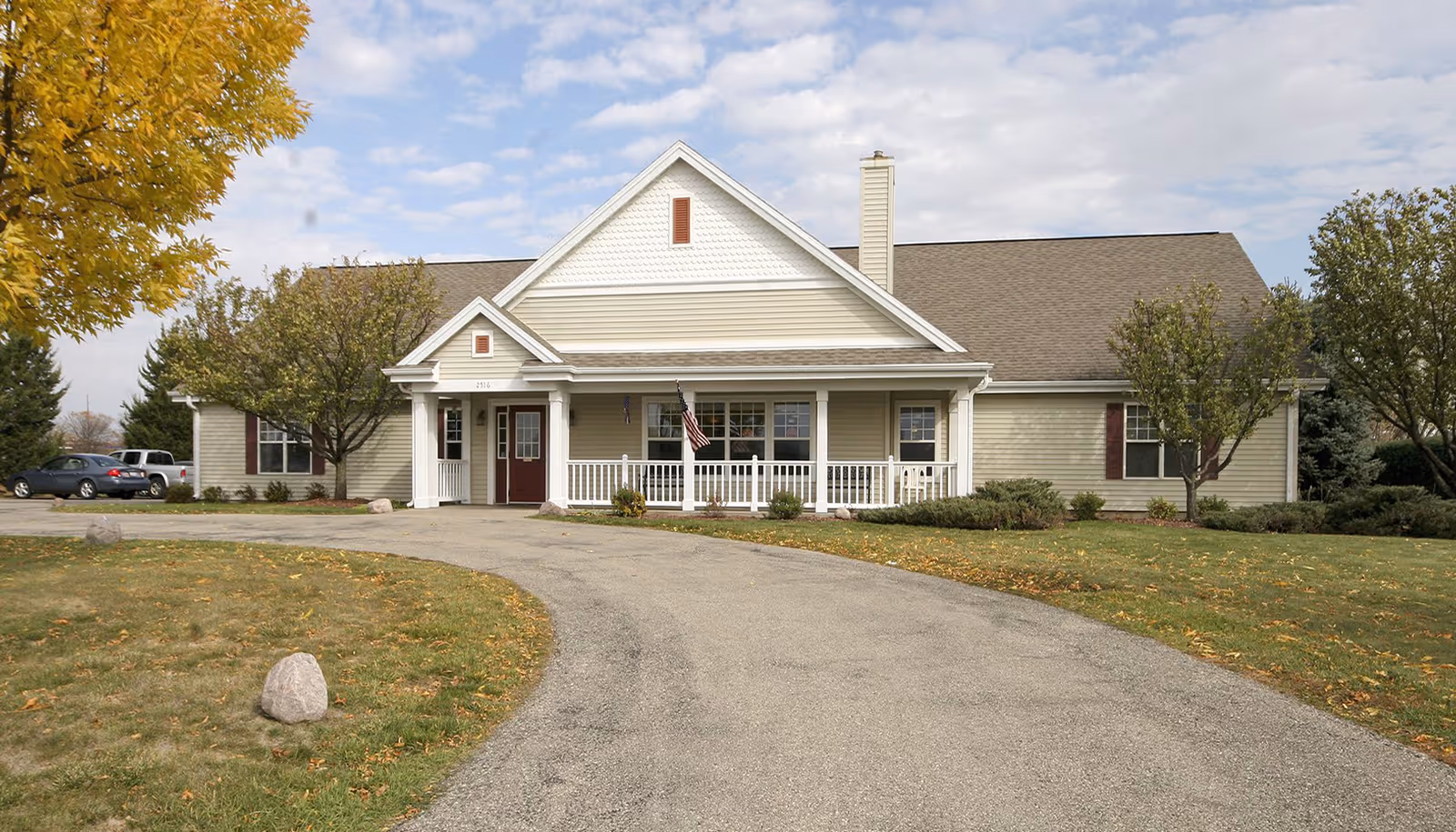 Front exterior of a one-story assisted living building with a curved driveway, a porch with white railing and an American flag, and surrounding lawn and trees.
