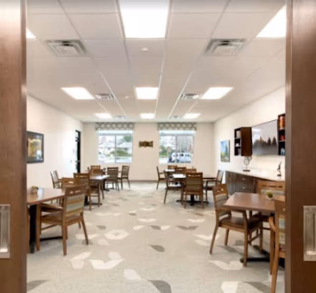 A bright dining room with multiple wooden tables and chairs arranged neatly. The room has large windows at the far end letting in natural light, a patterned carpet, and a wall-mounted television with cabinets underneath on the right side.