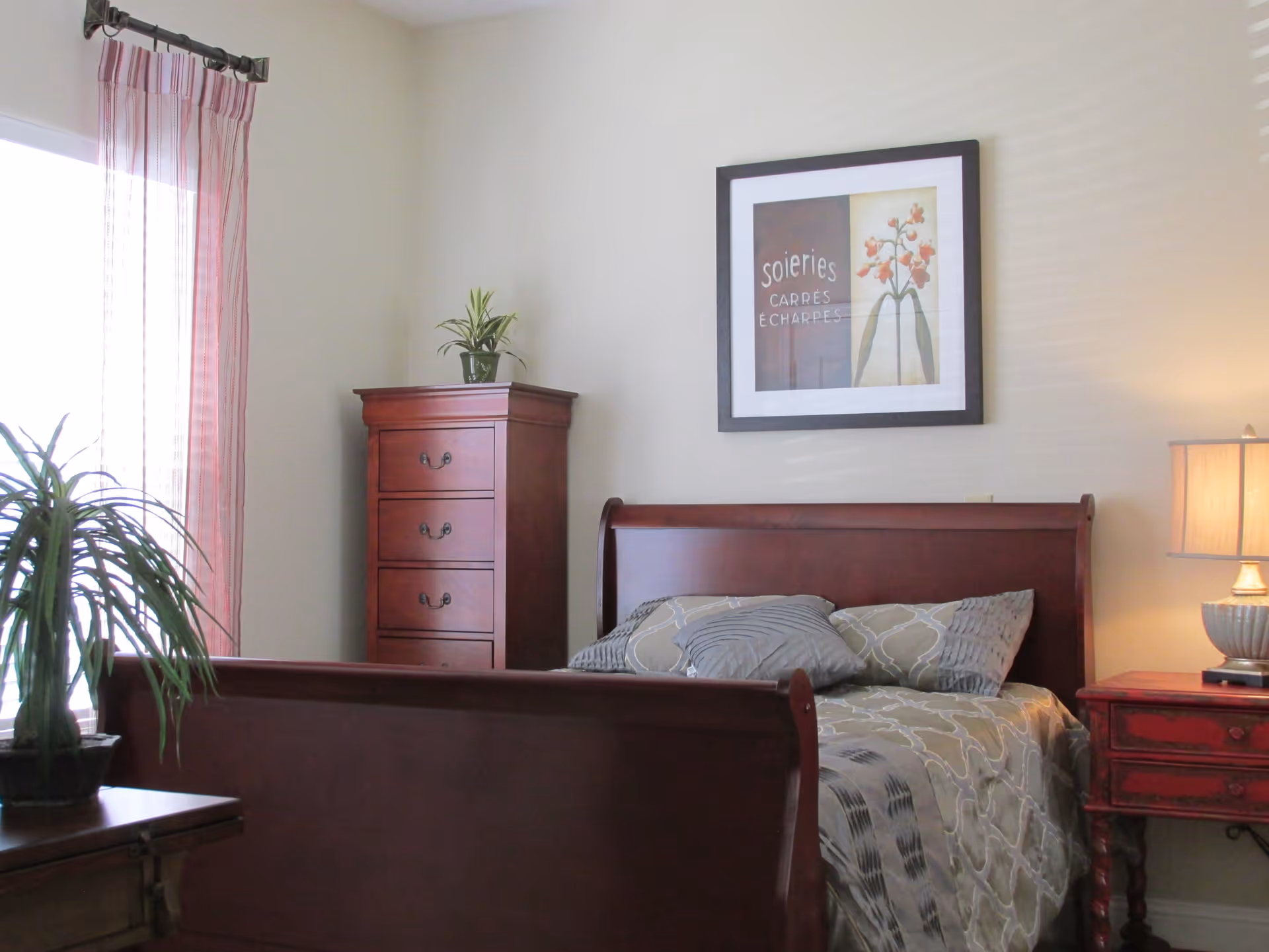 A bedroom with a wooden bed frame, a bed with patterned bedding and pillows, a wooden chest of drawers with a plant on top, a wooden nightstand with a lamp, a framed floral artwork on the wall, and a window with striped curtains.