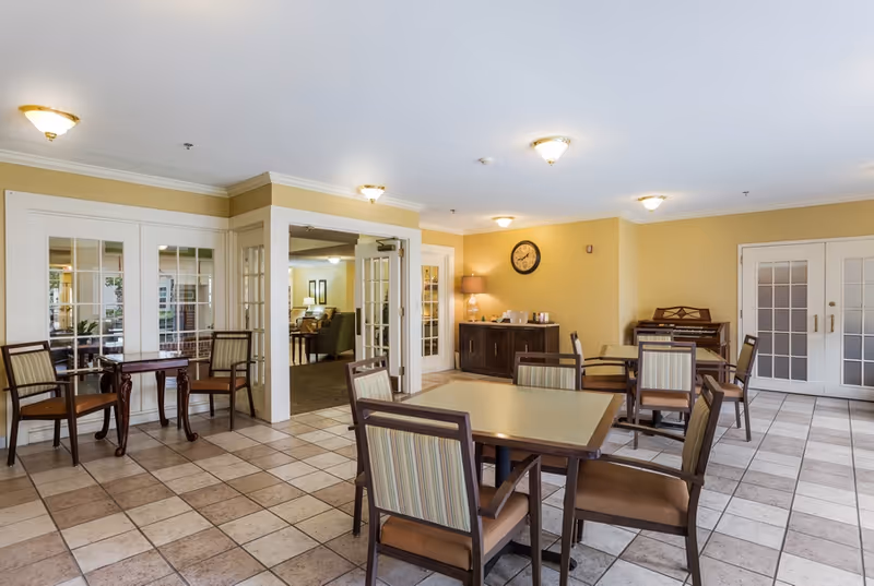 Bright communal dining room with several tables and chairs, tiled floor, French doors, and a sideboard with a clock and lamp.