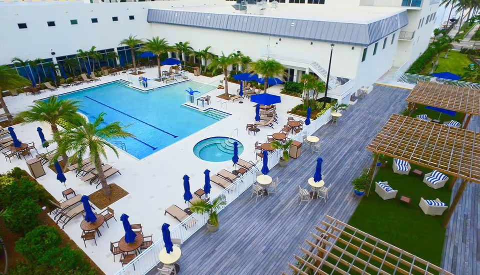 Aerial view of an outdoor pool and adjacent deck with lounge chairs, umbrellas, palm trees, and pergola-covered seating areas.