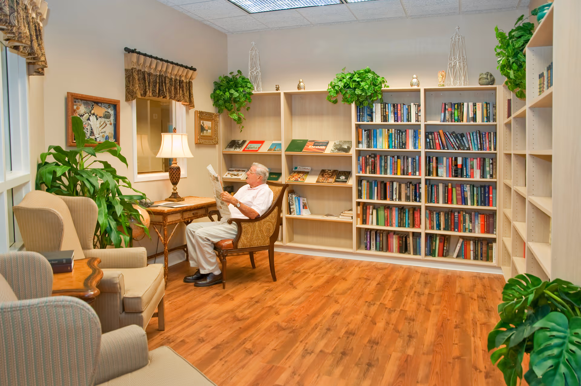 A senior man sitting in a chair reading a newspaper in a cozy library room with wooden floors, bookshelves filled with books, several green plants, and comfortable armchairs. A table lamp is on a small wooden table beside the man.