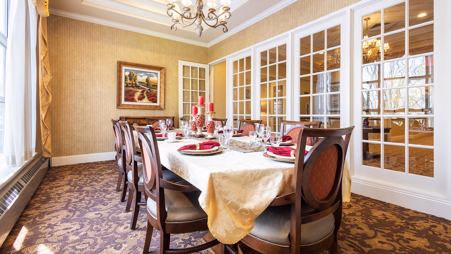 A formal dining room with a long table covered with a cream-colored tablecloth, set with plates, glasses, and red napkins. The room has patterned carpet, beige walls, a chandelier, and large windows with white-framed glass panels. A framed landscape painting hangs on the wall.