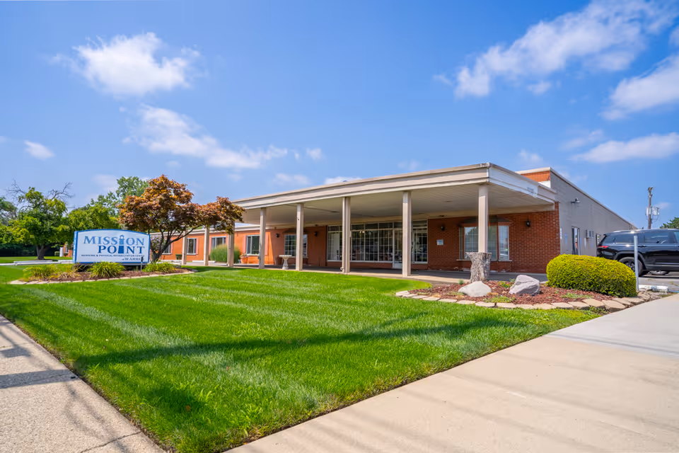 Exterior view of Mission Point Nursing & Physical Rehabilitation Center of Warren, showing a single-story brick building with a covered entrance, well-maintained green lawn, small trees, and a clear blue sky with some clouds.