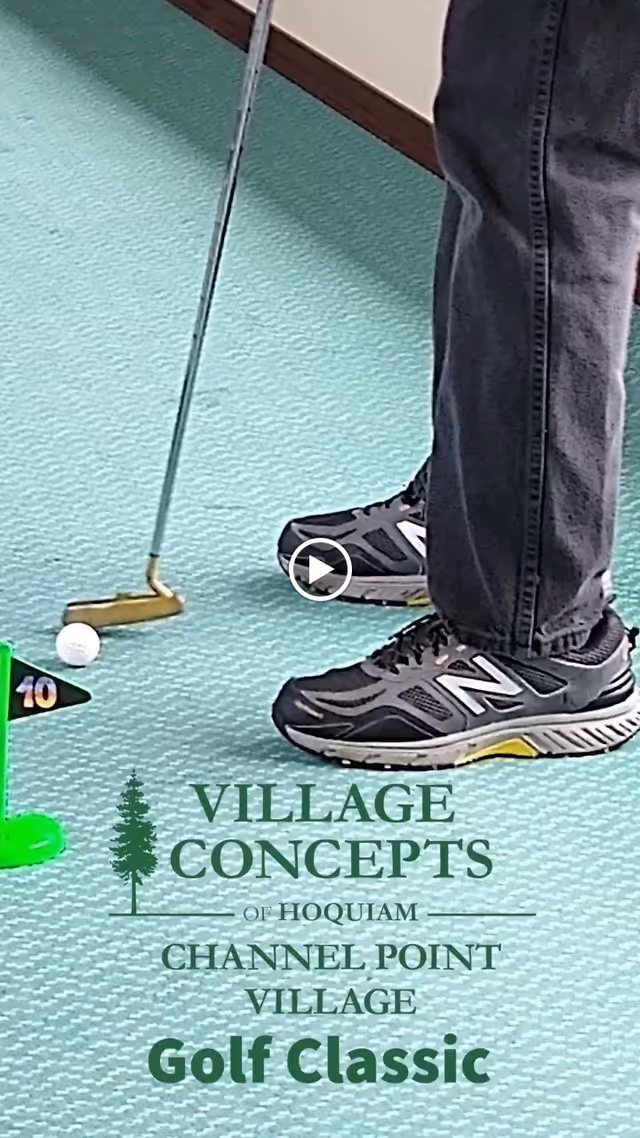 Close-up of a person wearing black New Balance sneakers and dark pants playing indoor mini-golf on a light blue carpet. A putter is positioned near a white golf ball next to a small green flag marked with the number 10.