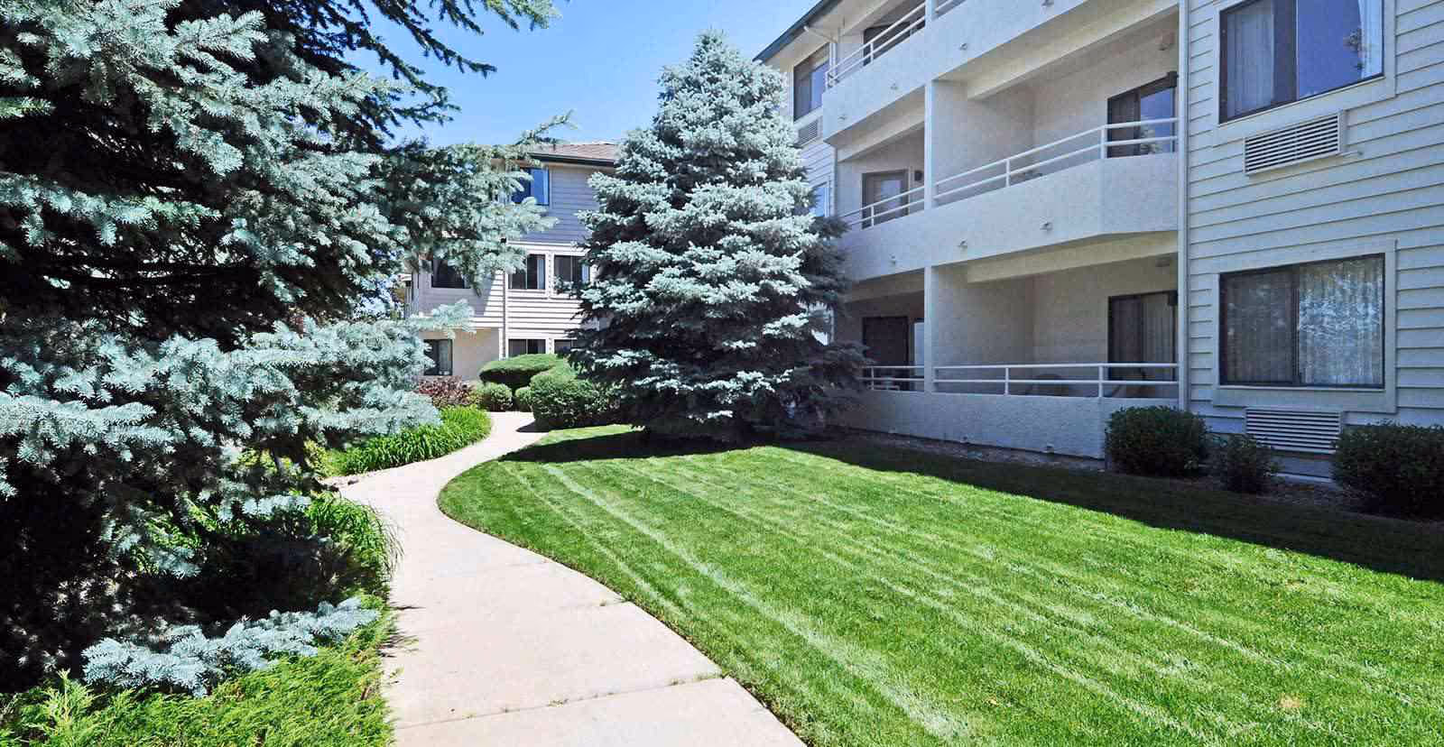 A landscaped courtyard with a curved sidewalk, green lawn, evergreen trees, and a multi-story building with balconies and windows.