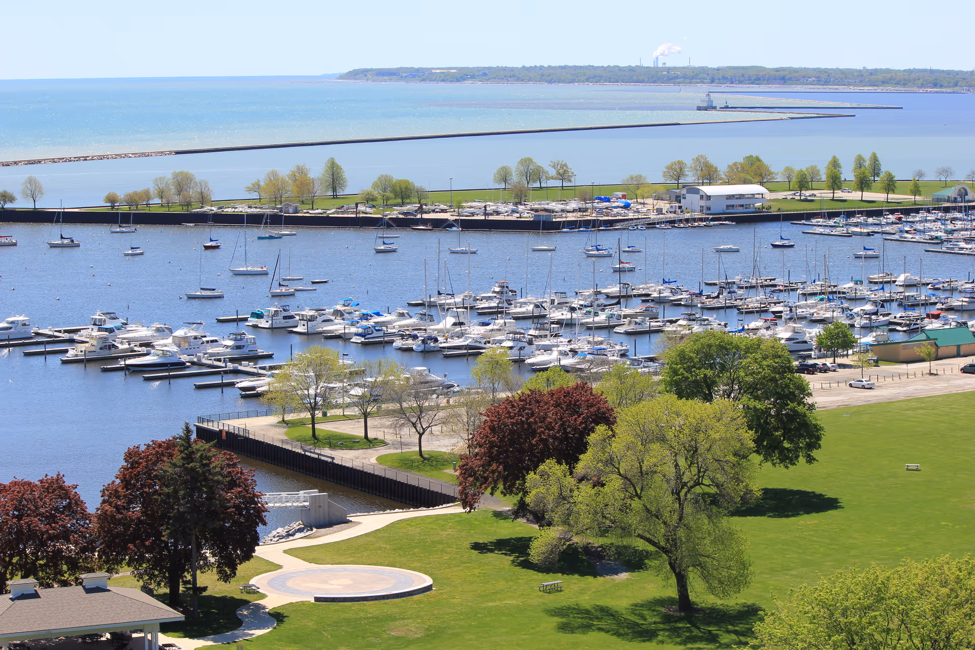 A scenic view of a marina filled with numerous boats docked in the water, surrounded by green trees and grassy areas. In the background, there is a large body of water extending to the horizon with a distant shoreline and some industrial structures visible.