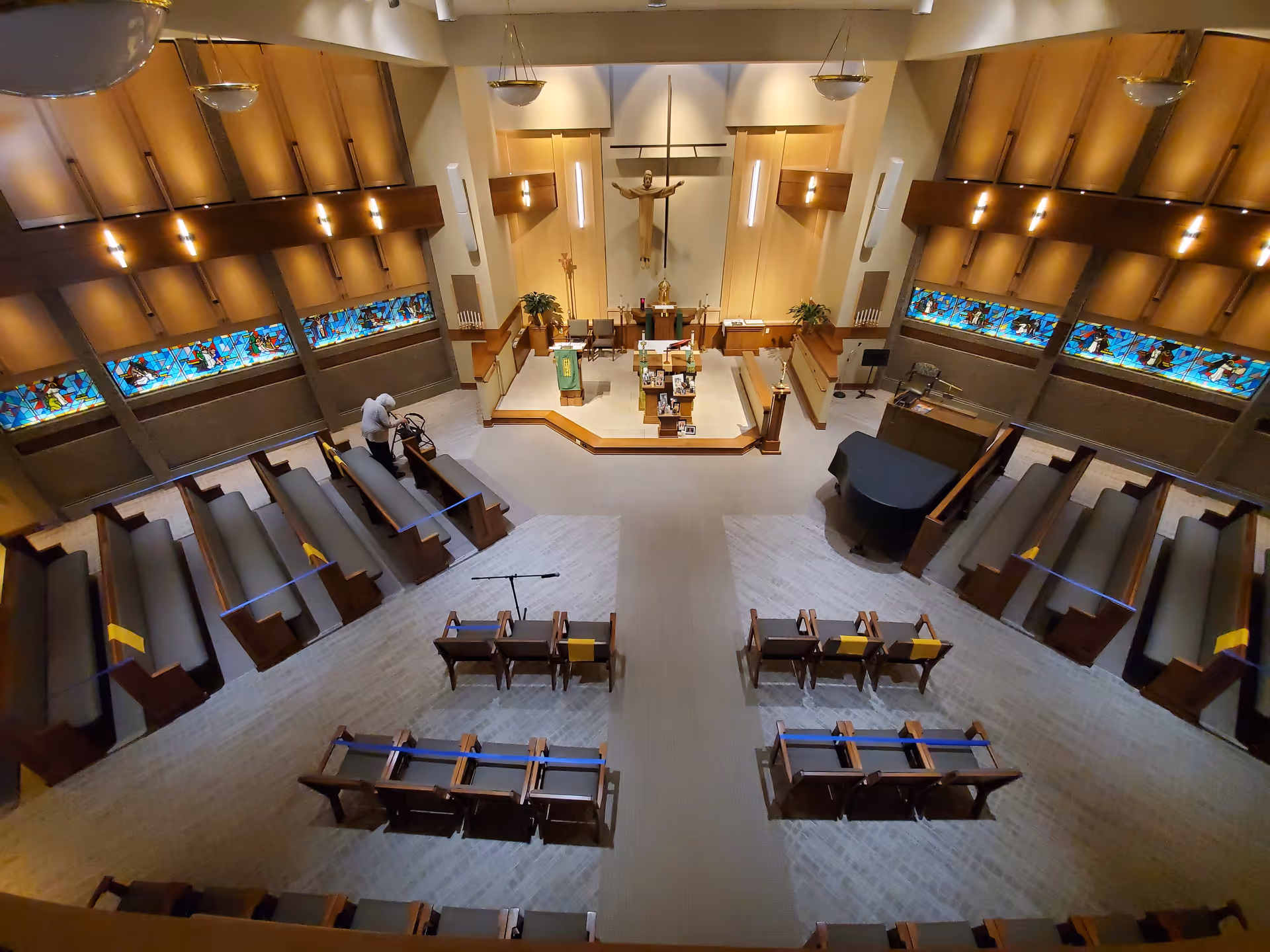 Interior view of a chapel with wooden pews arranged facing an altar. The altar area features a large crucifix on the wall, plants, and religious items. Stained glass windows run along the side walls. A person with a walker is seen near the pews on the left side. The lighting is warm and the ceiling has hanging light fixtures.