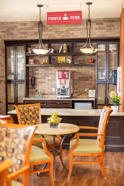 A cozy coffee station labeled "Power Perk" with a beverage machine, cabinets, and round tables with chairs in a communal area.