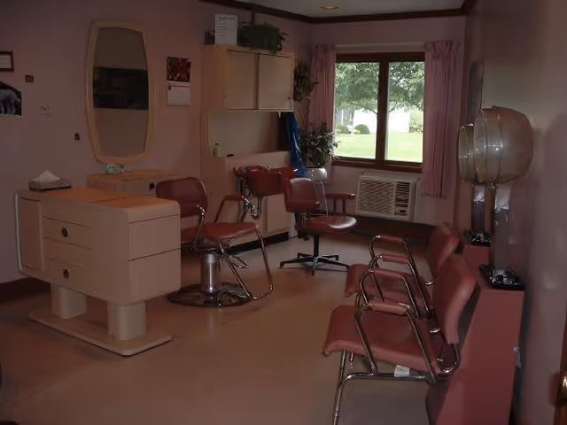 Interior of a salon area with multiple red salon chairs, hair dryers mounted on the wall, a vanity with a mirror, and a window with pink curtains overlooking a green lawn outside.