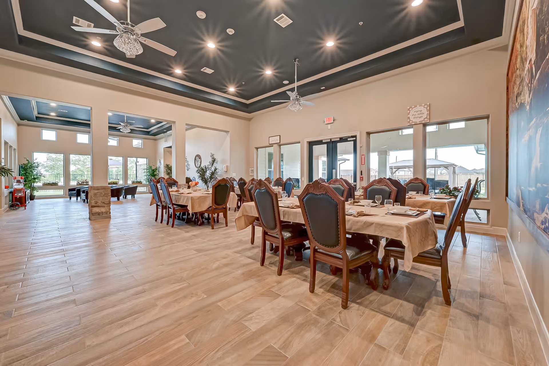 Spacious dining room in an assisted living facility with multiple tables covered with beige tablecloths, set with plates, glasses, and napkins. The room features high ceilings with recessed lighting and ceiling fans, large windows letting in natural light, and a view of an outdoor patio area.
