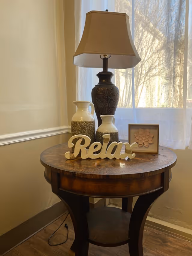 A round wooden side table with a decorative lamp, two ceramic pitchers, a framed flower artwork, and a wooden sign that says 'Relax' placed on top. The table is positioned near a window with sheer white curtains, allowing natural light to filter through.