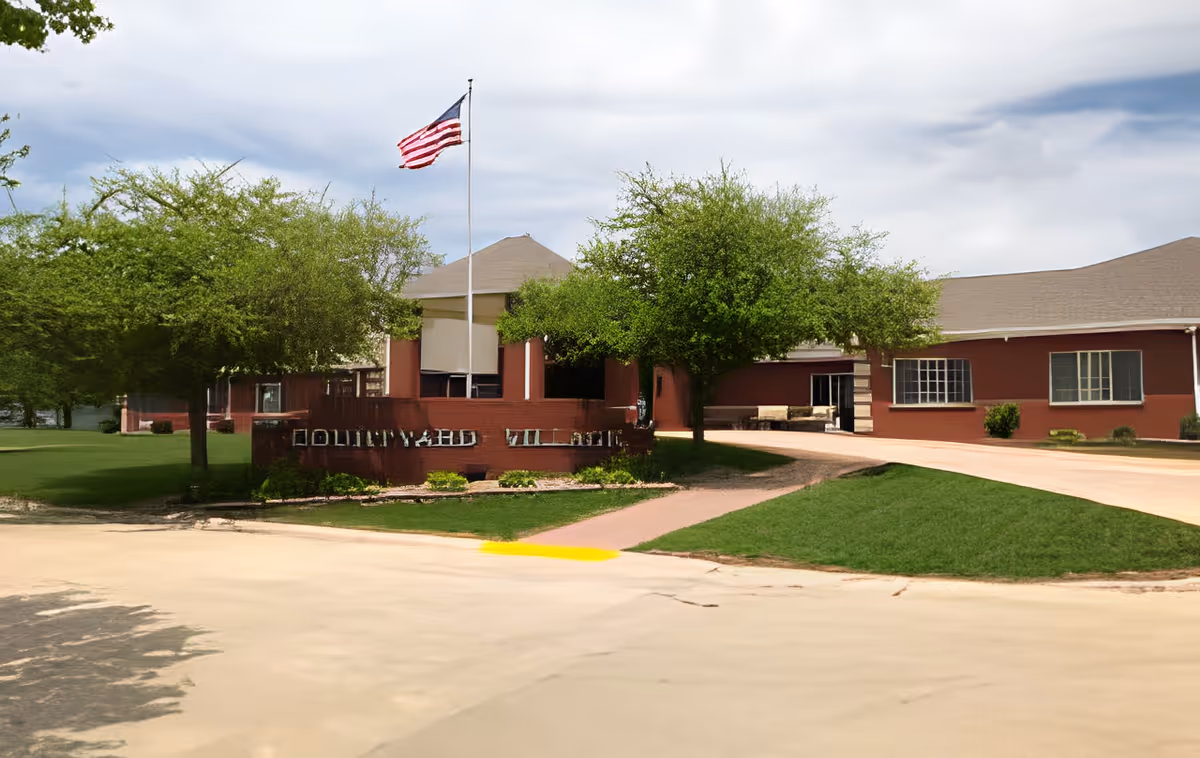 Exterior view of a single-story brick building with a driveway, green trees, and an American flag on a flagpole in front. The building has multiple windows and a sign in front partially obscured by shadows and trees.