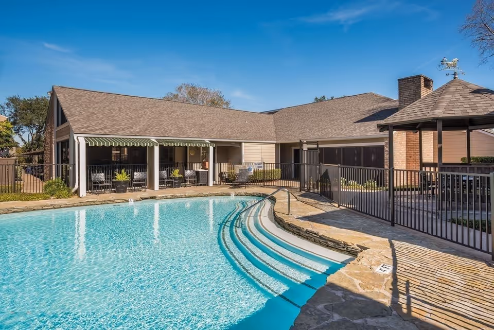 Outdoor swimming pool with clear blue water and curved steps leading into the pool. Surrounding the pool is a stone deck and black metal fencing. In the background, there is a single-story building with a sloped roof, a covered patio area with chairs and potted plants, and a gazebo with a weather vane on top. The sky is clear and blue.