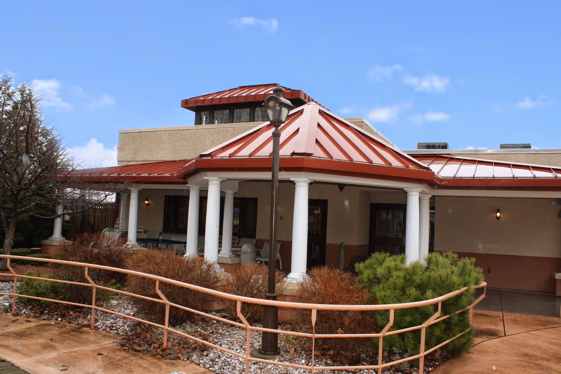 Entrance of a single-story building with a red metal roof, white columns, a lamp post, and a landscaped ramp.