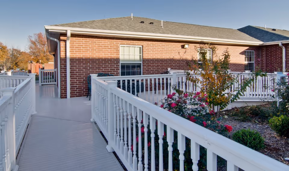 A white-railed outdoor ramp and walkway leading to a red brick senior living building with landscaped flower beds.