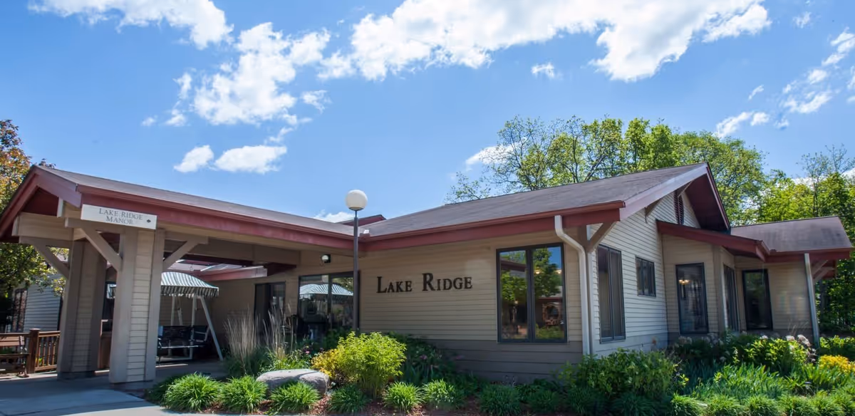 Exterior view of a single-story building with beige siding and a red roof trim under a blue sky with scattered clouds. The building has a sign that reads 'Lake Ridge' and a covered entrance area with a sign pointing to 'Lake Ridge Manor'. There are green shrubs and plants around the building.