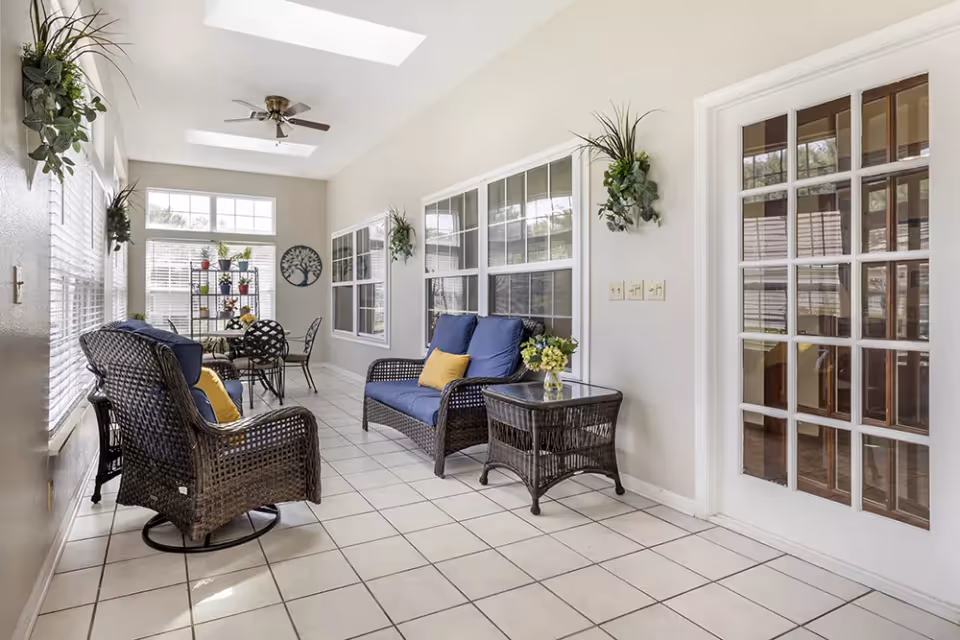 Bright sunroom with white tiled floor and beige walls featuring wicker furniture with blue cushions and yellow pillows, a glass-top table with black metal chairs, several potted plants on a black metal shelf, wall-mounted plants, and large windows allowing natural light to fill the space.