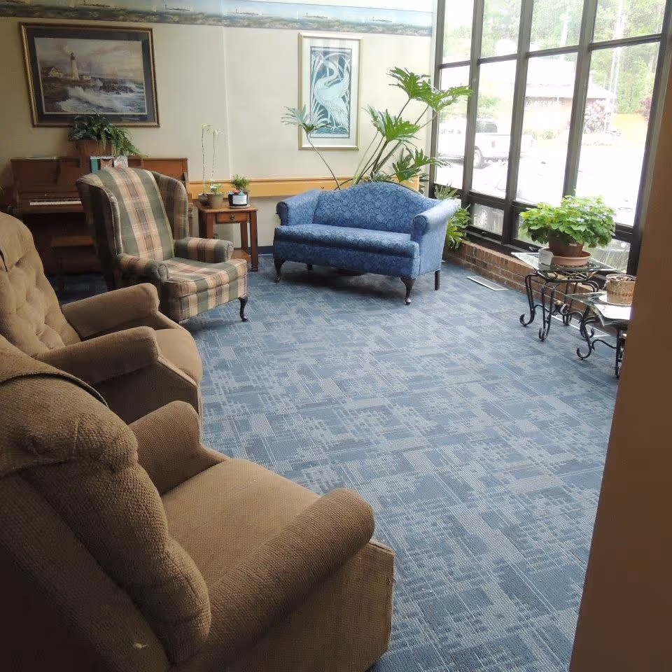 A cozy sitting area with a beige recliner, a plaid armchair, and a blue upholstered loveseat arranged on a blue patterned carpet. There are several potted plants on small tables near large windows letting in natural light. A piano and framed artwork are visible on the walls.