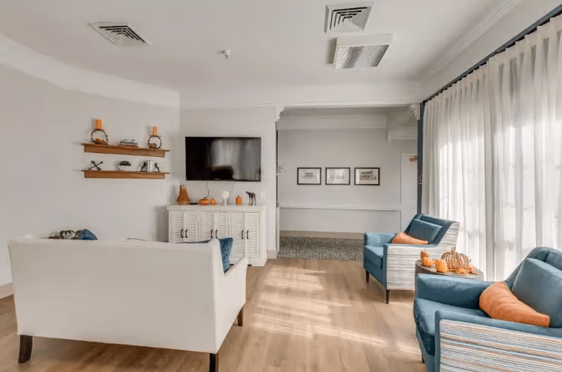 A bright living room with light wood flooring, a white sofa facing a wall-mounted flat screen TV above a white cabinet decorated with small pumpkins and vases. Two blue armchairs with orange cushions are positioned near large windows covered with sheer white curtains. Wooden shelves with decorative items are mounted on the wall to the left.