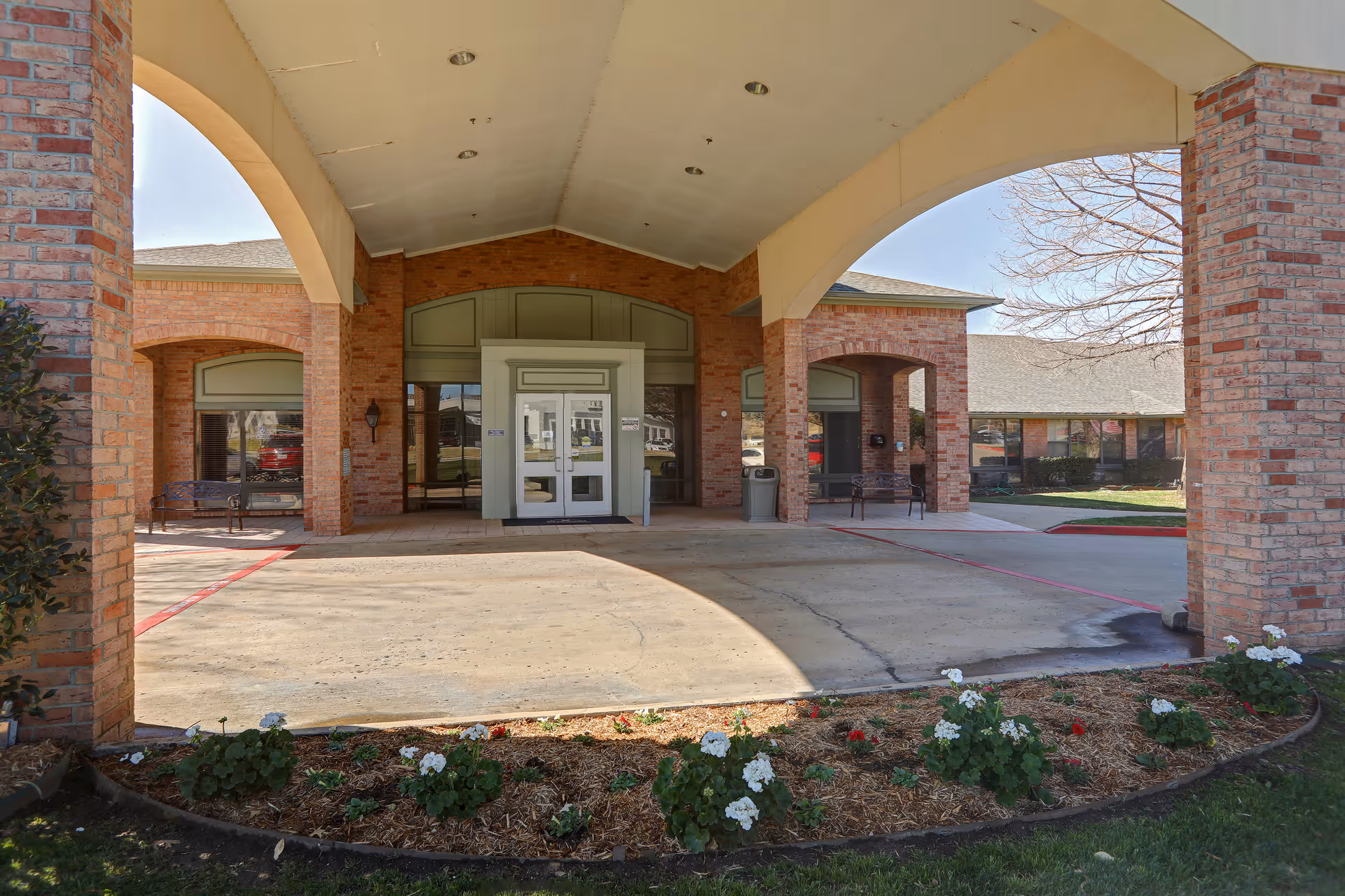 Entrance area of a brick building with a covered driveway, white double doors, benches on either side, and a small garden bed with flowers in the foreground.
