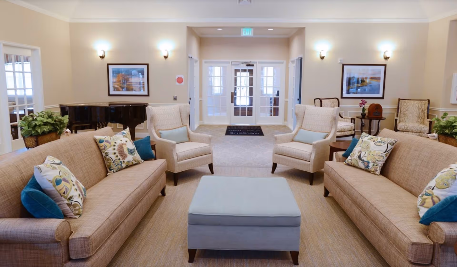 A spacious and well-lit living room area with two beige sofas adorned with colorful patterned and solid blue pillows, two beige armchairs with light blue cushions, and a light blue ottoman in the center. The room features beige walls with framed artwork, wall sconces, potted plants, and a black grand piano in the corner. Double glass doors lead to another area, and the overall decor is warm and inviting.