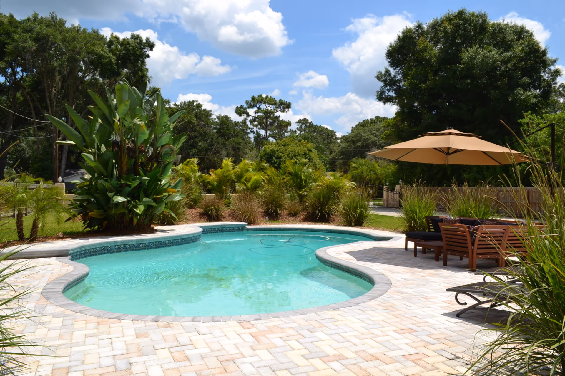 Outdoor swimming pool with clear blue water surrounded by a paved patio area. There are green plants and trees around the pool, and a seating area with wooden furniture and a large beige umbrella on the right side. The sky is partly cloudy with blue patches.