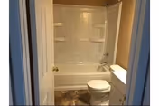 A small bathroom featuring a white bathtub with built-in shelves, a white toilet, and a white vanity with a sink. The floor has a patterned tile design, and the walls are painted beige.