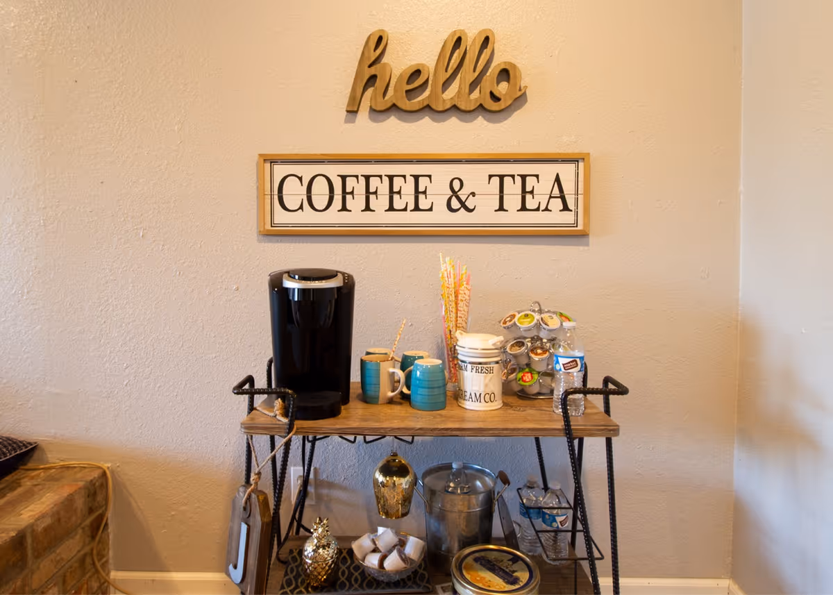 A coffee and tea station with a black coffee maker, three blue mugs, a container with colorful straws, a carousel of coffee pods, and various containers on a two-tier wooden and metal cart. Above the station, there are two wall decorations: a wooden cutout word 'hello' and a framed sign that reads 'COFFEE & TEA'.