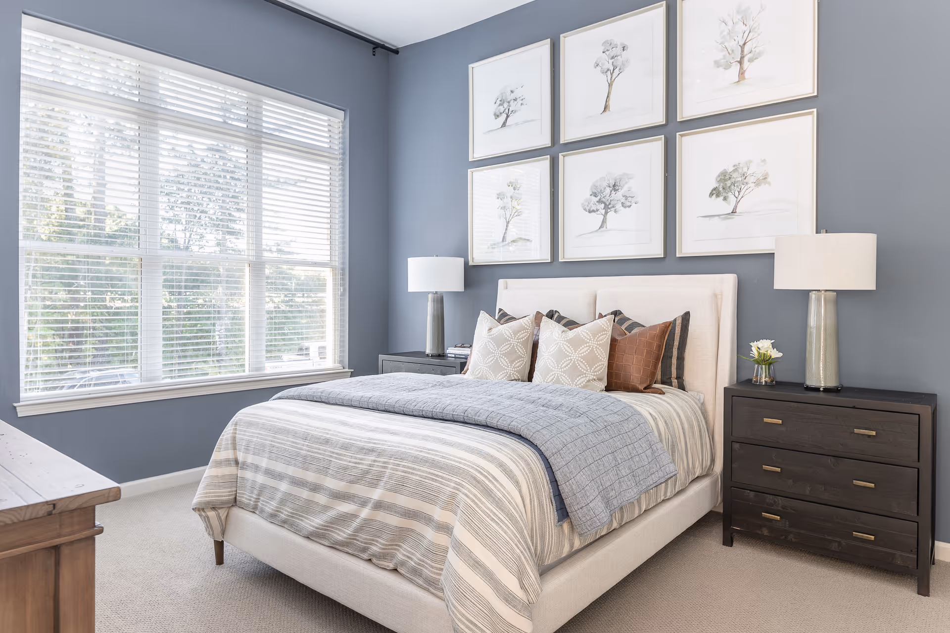 A neatly made bed with striped bedding and multiple pillows in a bedroom with blue walls. There are six framed pictures of trees arranged in two rows above the bed. Two dark wooden nightstands with lamps and a small flower vase flank the bed. A large window with white blinds lets in natural light.