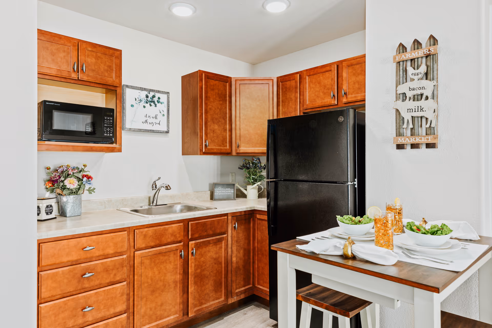 A compact kitchen with wooden cabinets, a black refrigerator, a microwave, and a stainless steel sink. There is a small dining table set for two with bowls of salad and glasses of iced tea. The walls are decorated with a framed quote and a wooden sign that reads 'Farmers Market eggs, bacon, milk.'