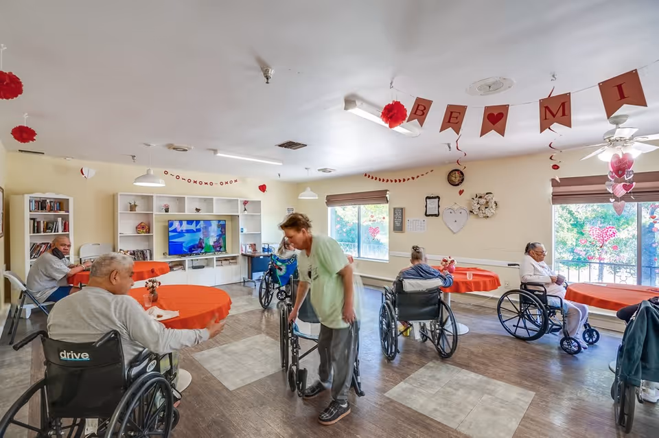 A common room in a senior living facility decorated with red and pink Valentine-themed decorations including banners and hanging ornaments. Several elderly residents in wheelchairs are seated around tables covered with red tablecloths. A caregiver is assisting one of the residents. The room has large windows letting in natural light, a TV on a white shelving unit, and bookshelves in the background.