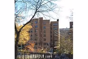 View of a multi-story brick apartment building surrounded by trees with some autumn foliage, seen from behind a black metal fence on a clear day.