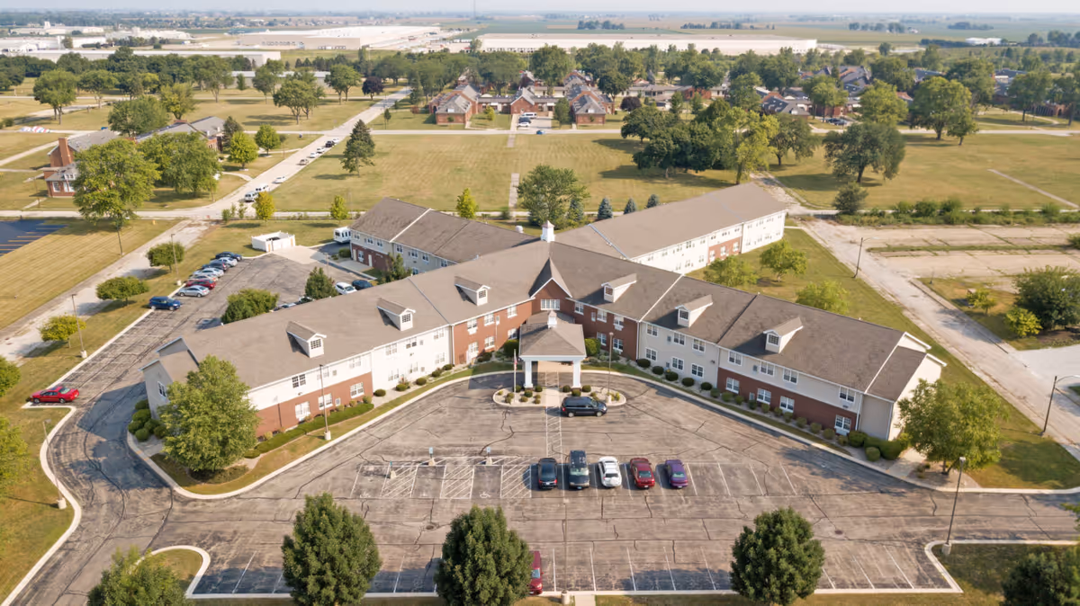 Aerial view of a curved two-story senior living building with a front entrance, parking lot, and surrounding lawns and trees.