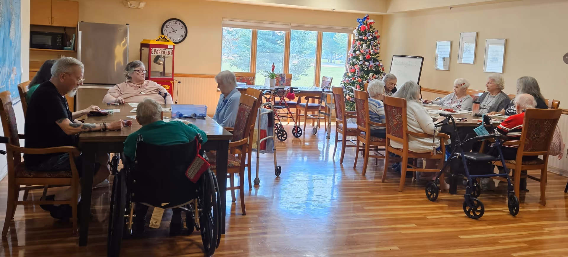 Residents seated around two large tables in a bright communal dining/activity room with wood floors, a Christmas tree, and windows.