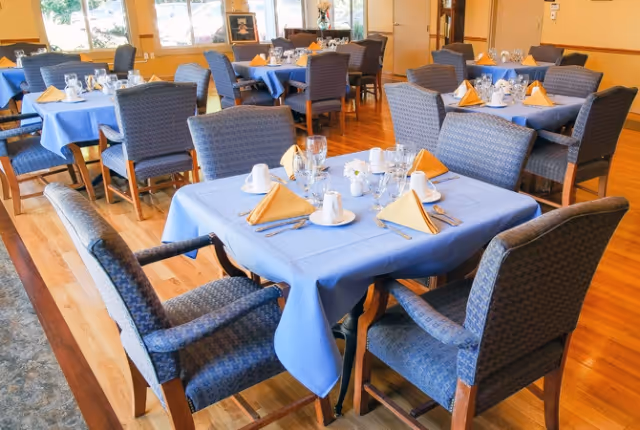 A dining room with multiple tables covered in blue tablecloths, each set with folded yellow napkins, white cups, glasses, and silverware. The chairs around the tables are upholstered in a blue patterned fabric. Large windows allow natural light to fill the room, and the floor is wooden.