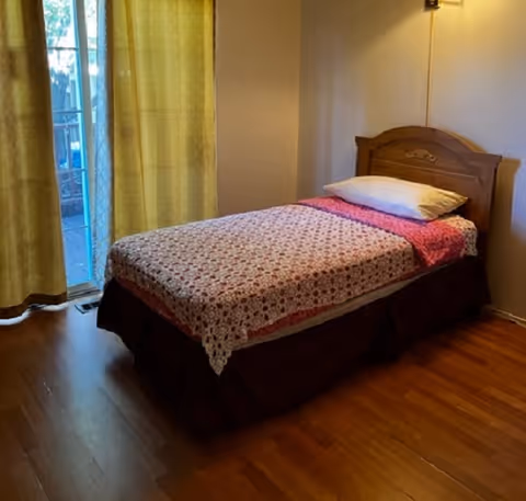 A simple bedroom with a single bed covered with a patterned bedspread and a pillow. The room has wooden flooring and light-colored walls. There is a window or glass door with yellow curtains allowing natural light to enter.