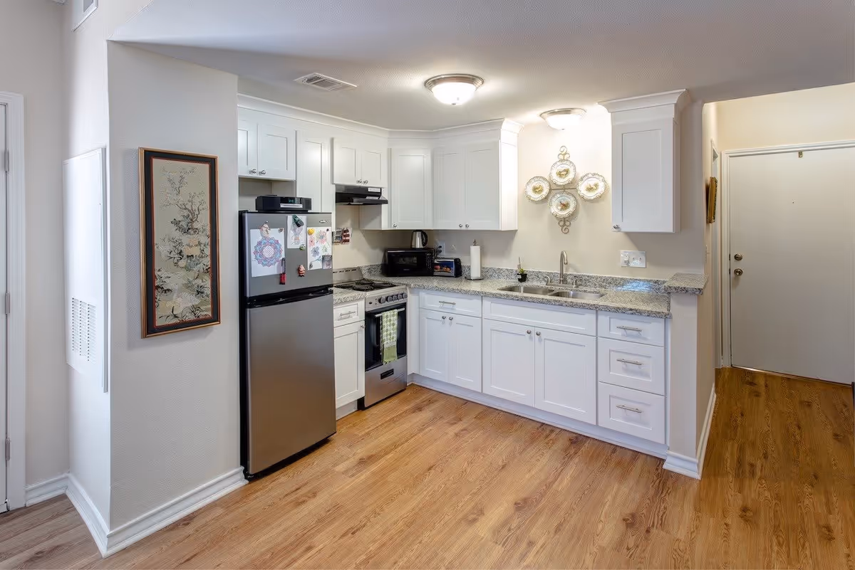 A modern kitchen with white cabinets, granite countertops, and wood flooring. The kitchen features a stainless steel refrigerator with magnets and drawings, a stove with an oven, a microwave, a double sink, and decorative plates mounted on the wall. The space is well-lit with ceiling lights and has a clean, organized appearance.