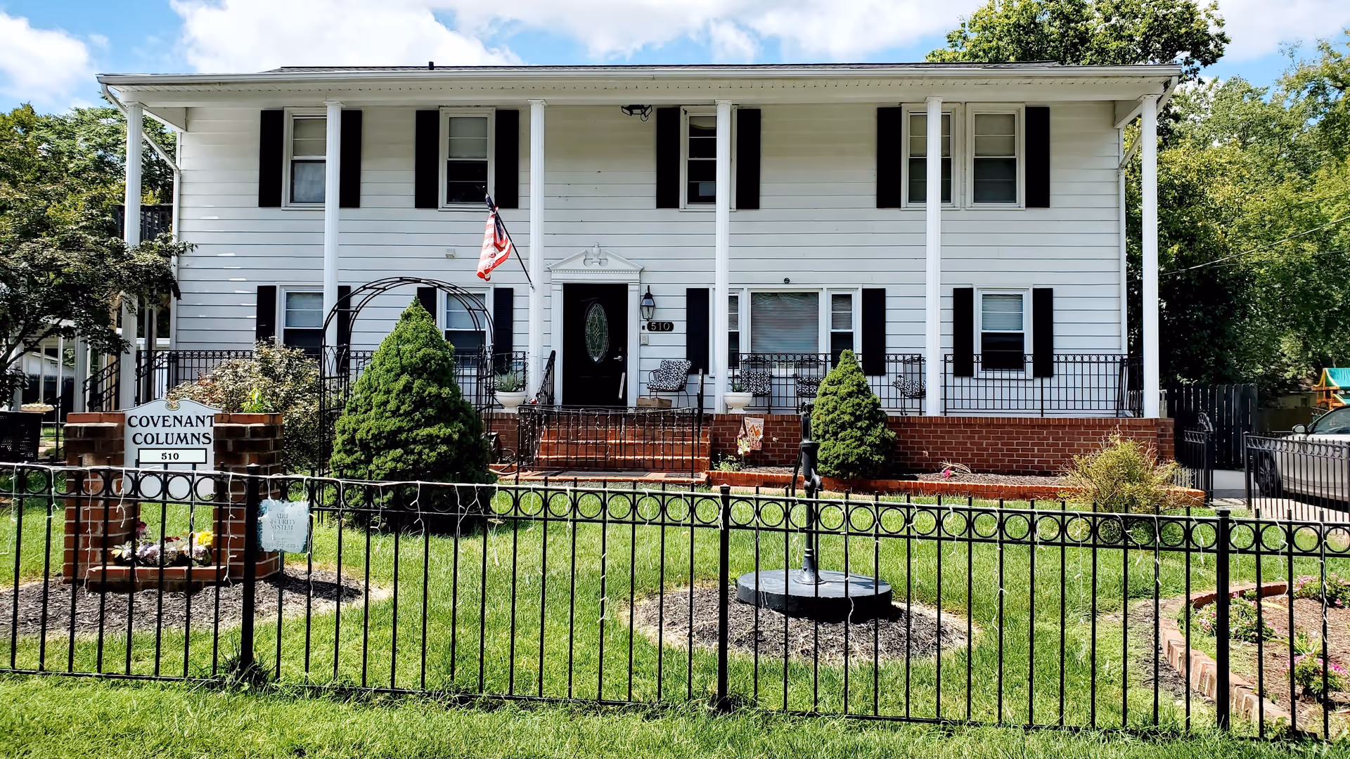 Front exterior view of a two-story white building with black shutters and a black front door, surrounded by a black metal fence. There are two small evergreen trees and a garden area in the front yard. A sign near the entrance reads 'Covenant Columns 510'. An American flag is mounted near the front door.