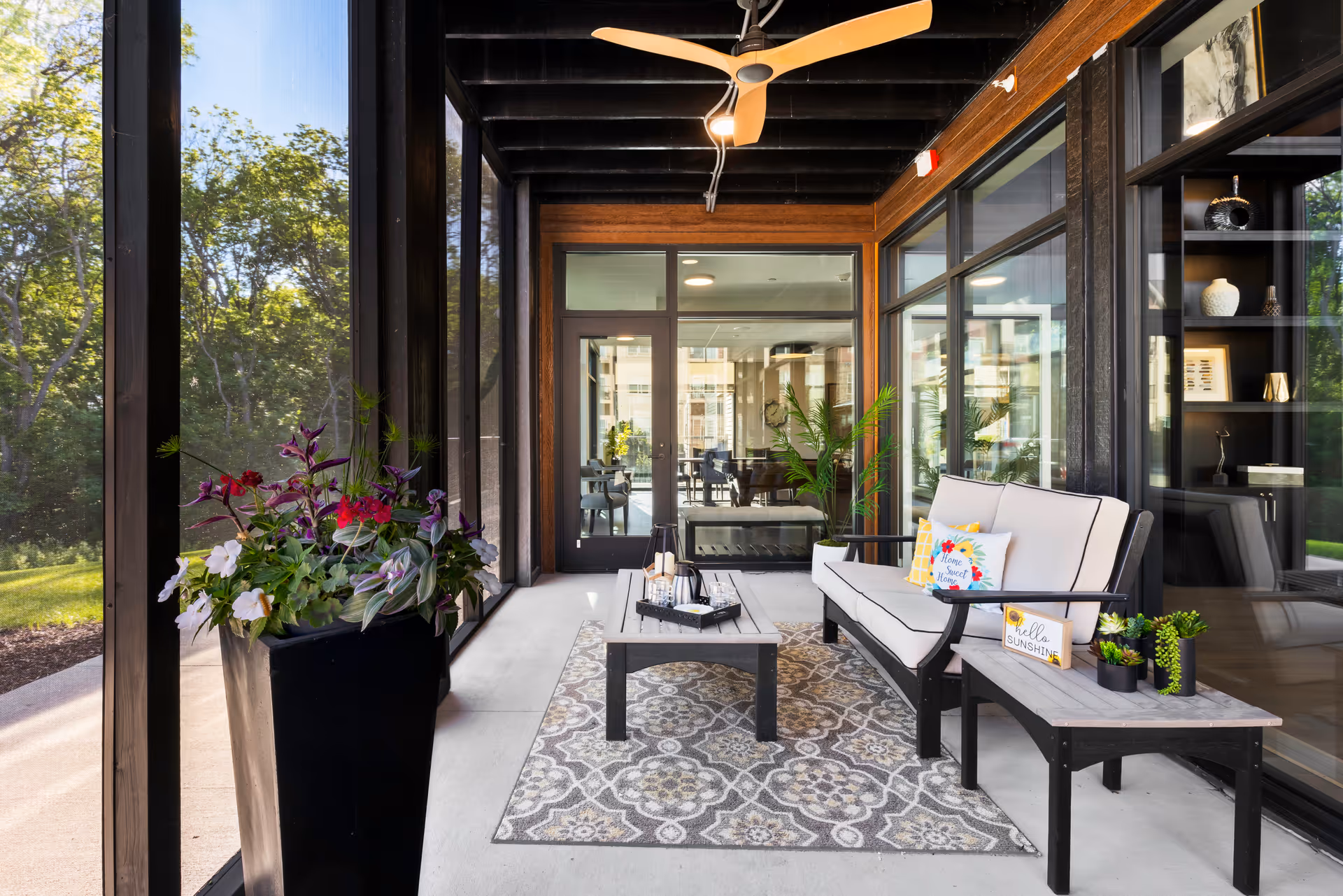 Screened outdoor lounge with a sofa, coffee table, rug, potted plants, and a ceiling fan adjacent to a building.