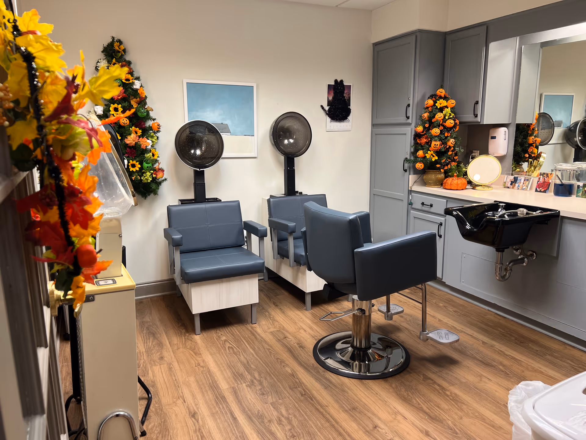 Interior of a hair salon area with two gray hair drying chairs and one styling chair in front of a black sink. The room has wood flooring, gray cabinets, and a countertop with various hair products. The space is decorated with small Halloween-themed decorations including pumpkin and flower arrangements.