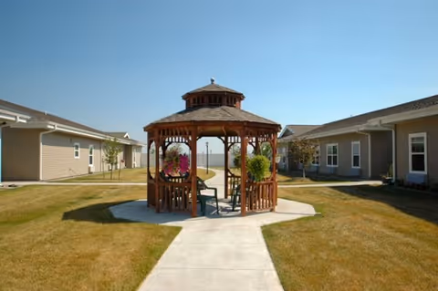 A wooden gazebo with hanging flower baskets situated in the center of a grassy courtyard surrounded by single-story beige buildings under a clear blue sky.