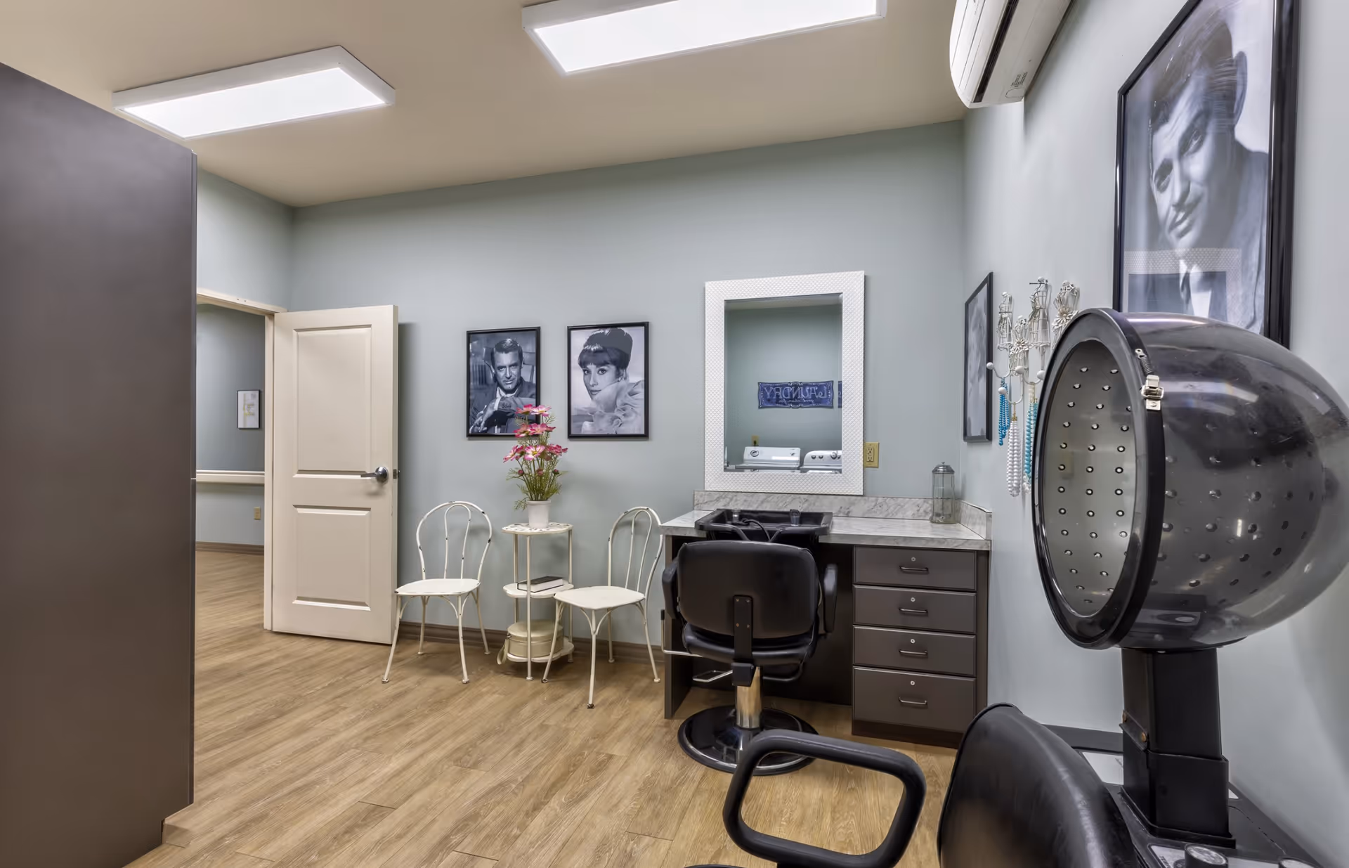 Interior of a hair salon room with a hair drying chair and hooded dryer, a styling chair in front of a counter with a mirror, two white metal chairs with a small round table holding a vase of flowers, and black and white framed portraits on the light blue walls.